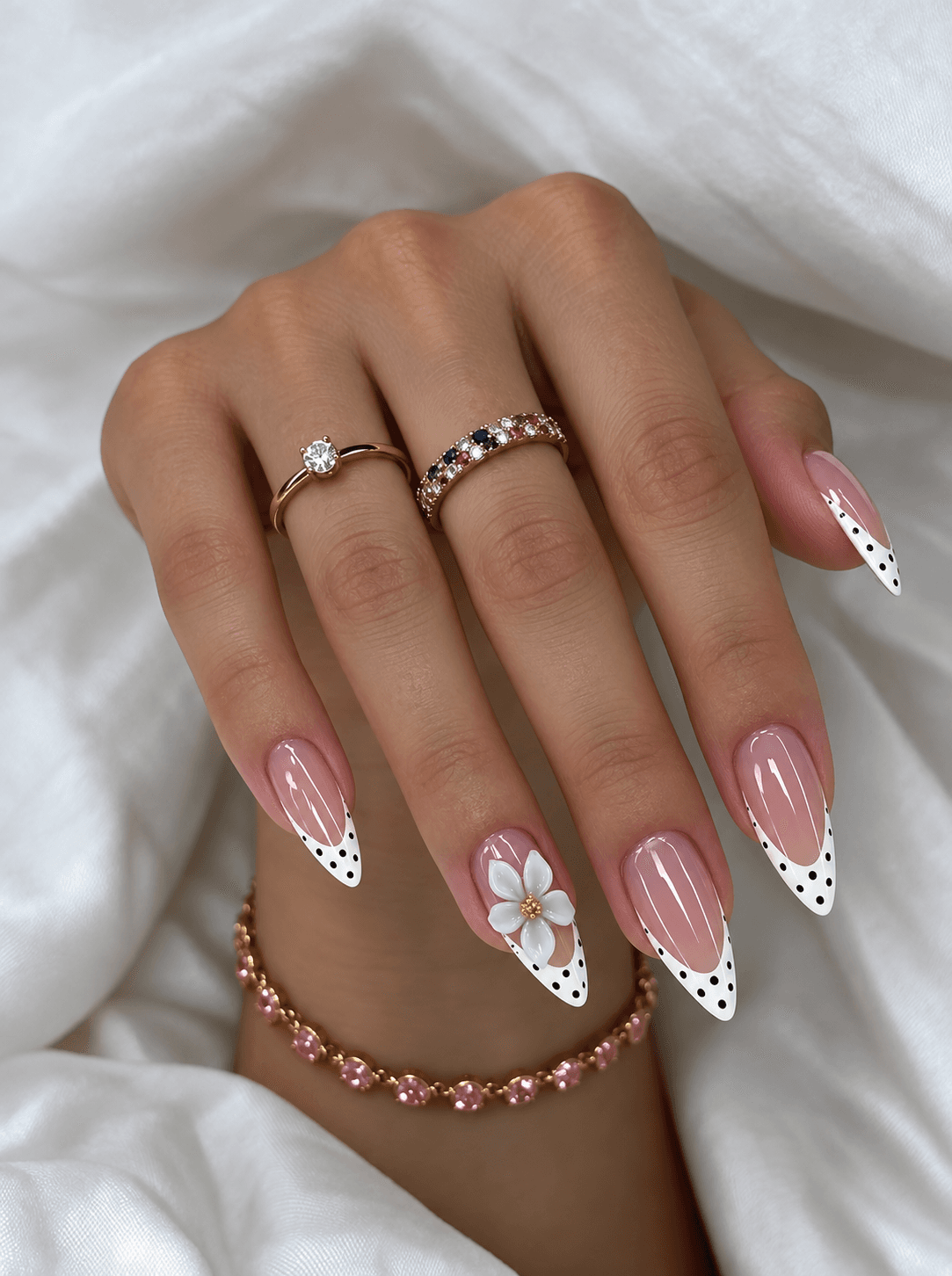 Nude nails with white polka-dot French tips and a raised white flower accent, worn with fine gold rings and a pink gemstone bracelet