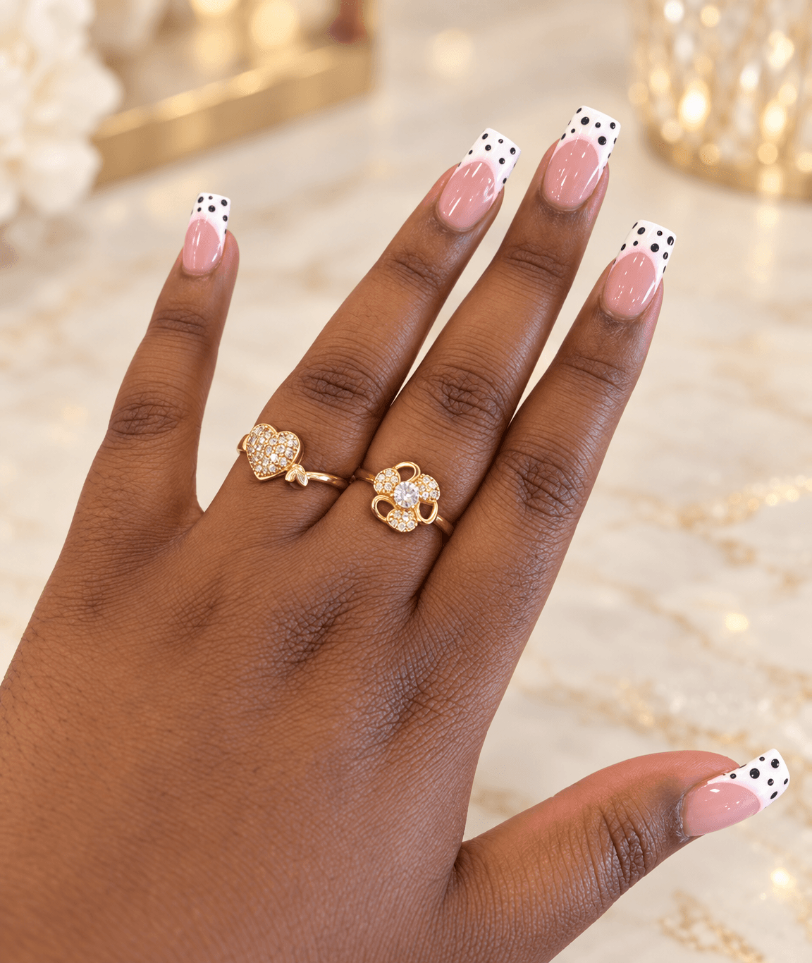 Square nails with white polka-dot French tips over a warm rose nude base, worn with diamante gold rings